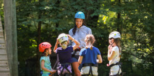 An adult in a blue helmet instructs four children in helmets and harnesses at an outdoor climbing or activity center on campus, surrounded by trees.