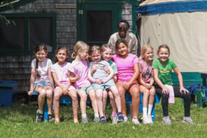 A group of eight young girls and one adult sit on a bench outdoors, smiling at the camera. Behind them is a rustic wooden building and a large tent.